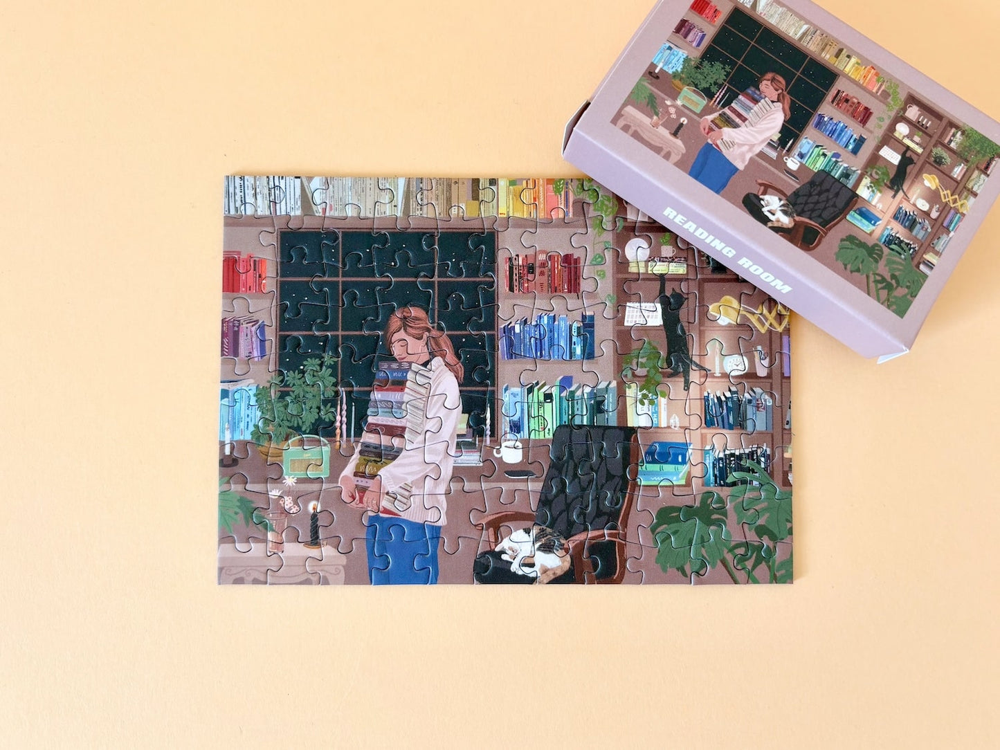 99-teiliges Minipuzzle für Erwachsene, das eine Frau mit einem Stapel Bücher in der Hand stehend vor ihrem gefüllten Bücherregal zeigt. Eine Katze schläft auf dem Stuhl neben ihr und eine andere klettert das Bücherregal hoch.