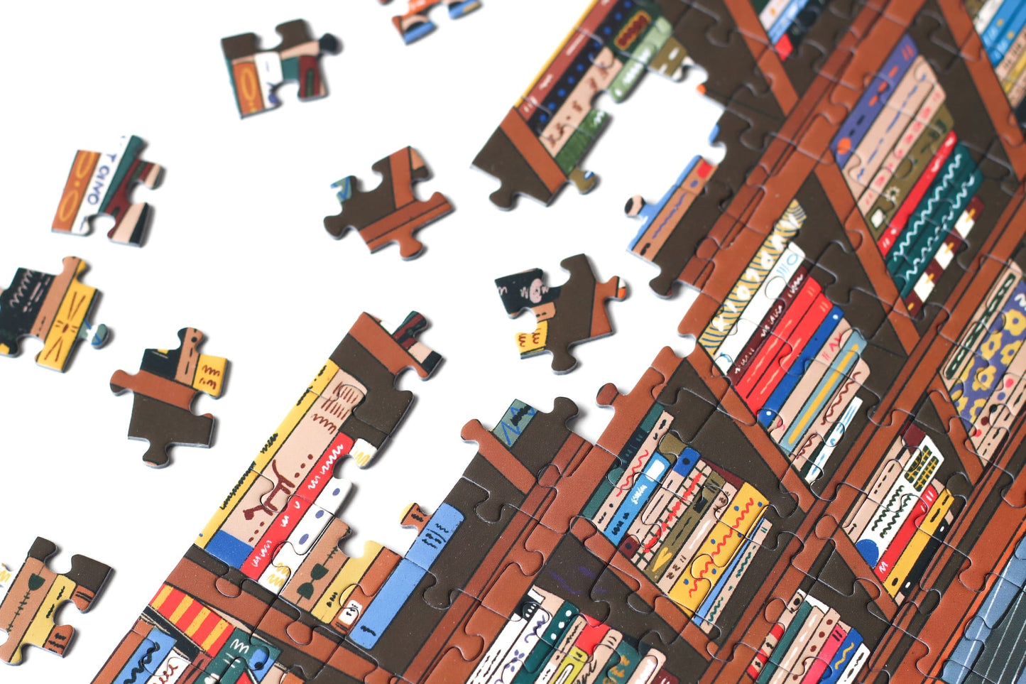 1000-Teile-Puzzle „Book Nook“ von Piecely – Illustration einer gemütlichen Bibliothek mit Bücherregalen, Lesenden und warmem Licht, auf brauner Verpackung.