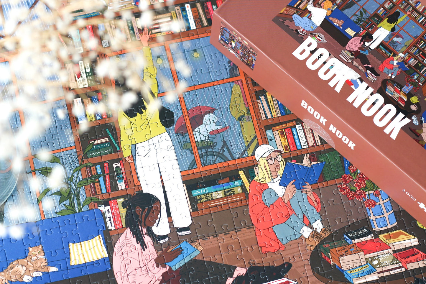 1000-Teile-Puzzle „Book Nook“ von Piecely – Illustration einer gemütlichen Bibliothek mit Bücherregalen, Lesenden und warmem Licht, auf brauner Verpackung.