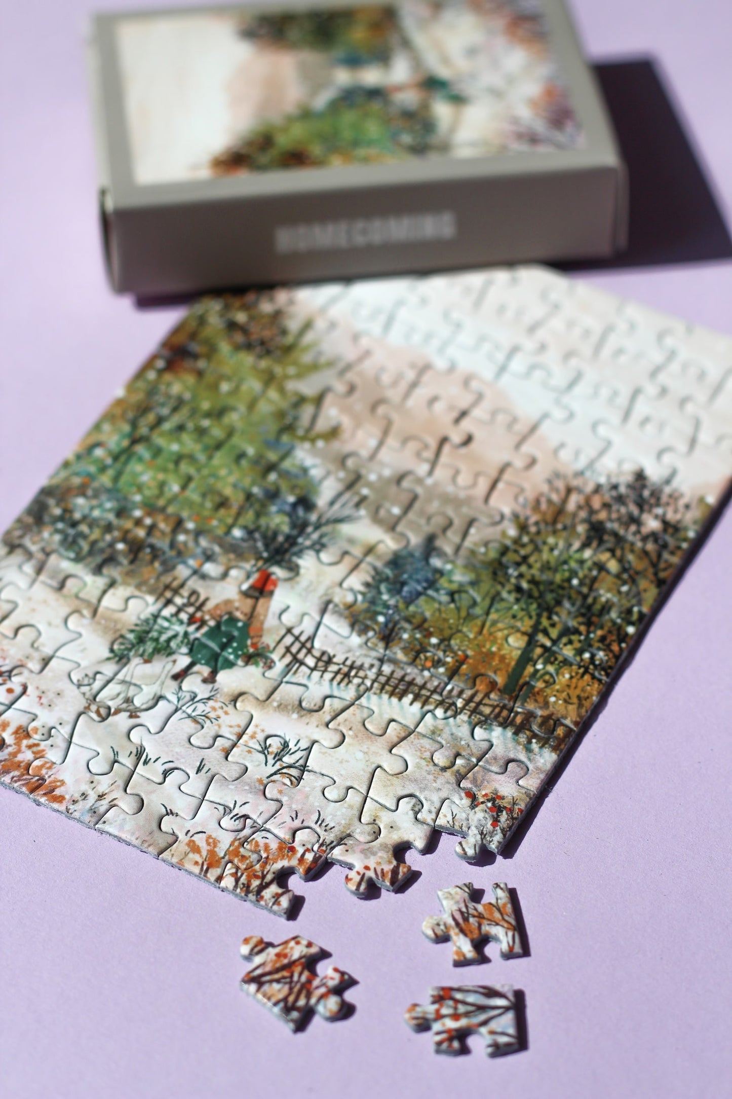 99-Teile-Mini-Puzzle „Homecoming“ von Piecely – Illustration einer winterlichen Landschaft mit Frau, zwei Gänsen und Weihnachtsbaum, auf grauer Verpackung