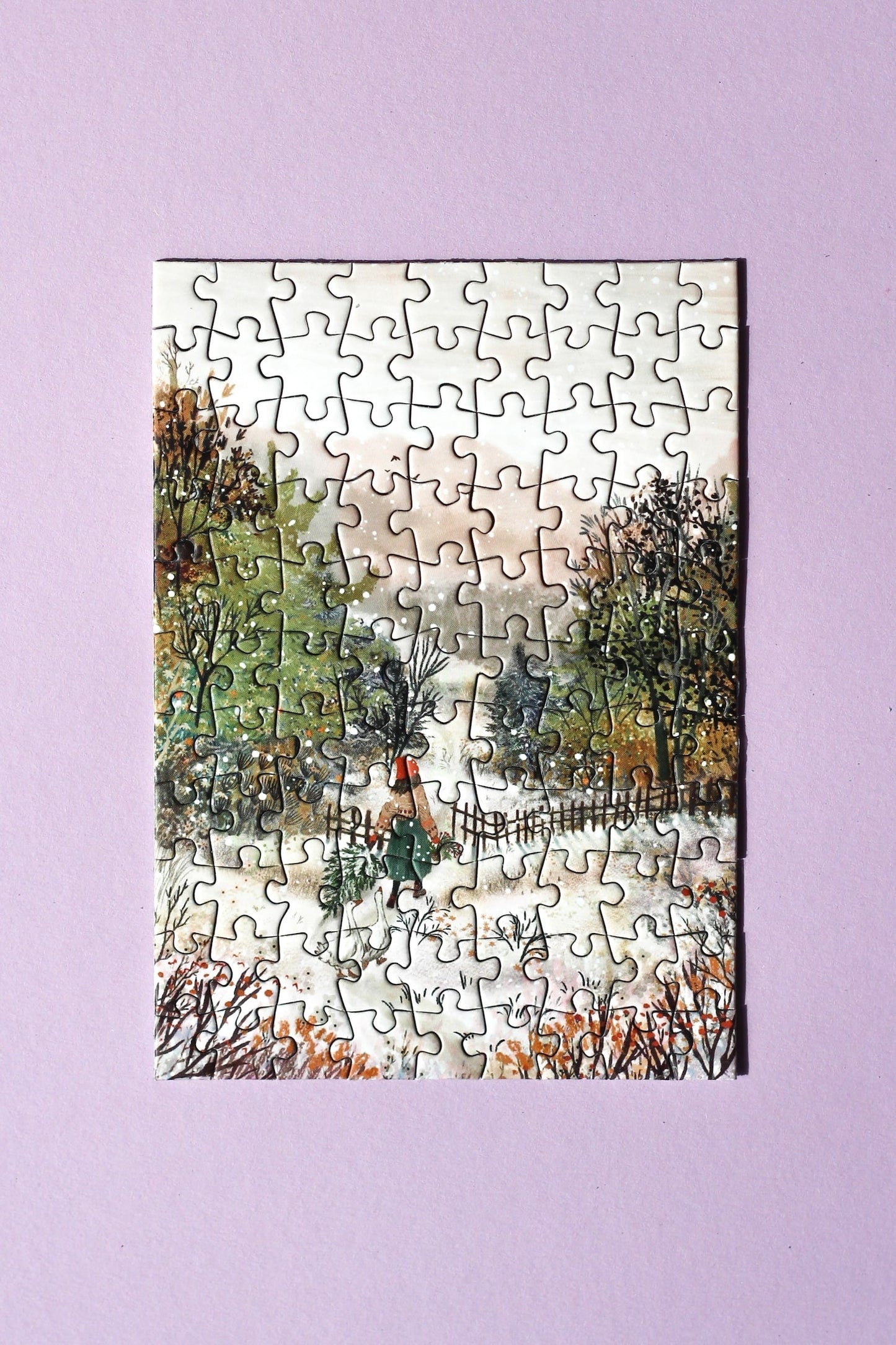 99-Teile-Mini-Puzzle „Homecoming“ von Piecely – Illustration einer winterlichen Landschaft mit Frau, zwei Gänsen und Weihnachtsbaum, auf grauer Verpackung