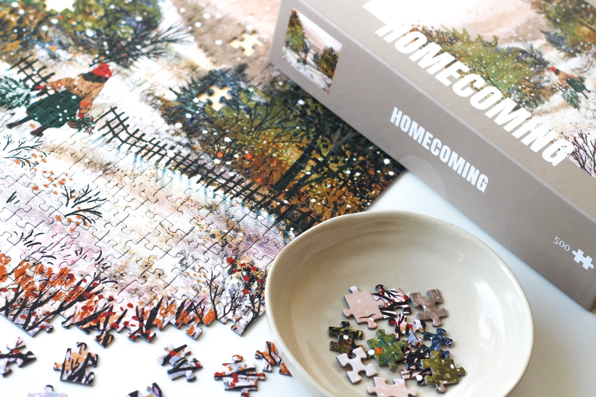 500-Teile-Puzzle „Homecoming“ von Piecely – Illustration einer winterlichen Landschaft mit Frau und zwei Gänsen auf einem verschneiten Weg, auf grauer Verpackung.