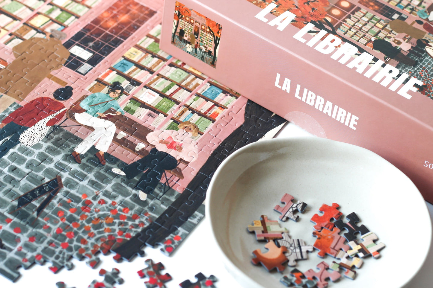 500-Teile-Puzzle „La Librairie“ von Piecely – Illustration einer herbstlichen Pariser Straßenszene vor einer kleinen Buchhandlung, auf rosa-brauner Verpackung.