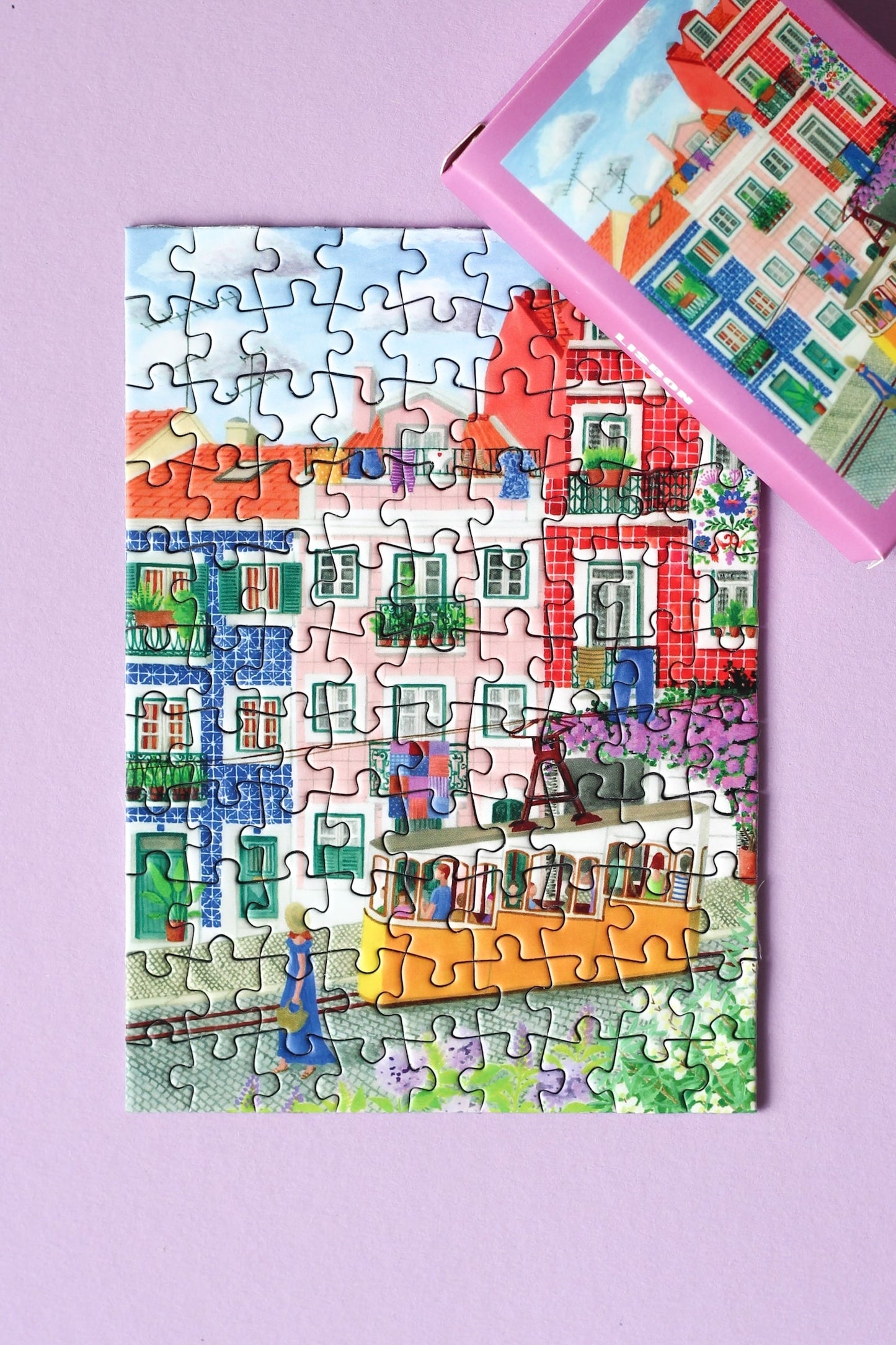 99-Teile-Mini-Puzzle „Lisbon“ von Piecely – Illustration einer farbenfrohen Straßenszene in Lissabon mit gelber Straßenbahn, auf pinker Verpackung.