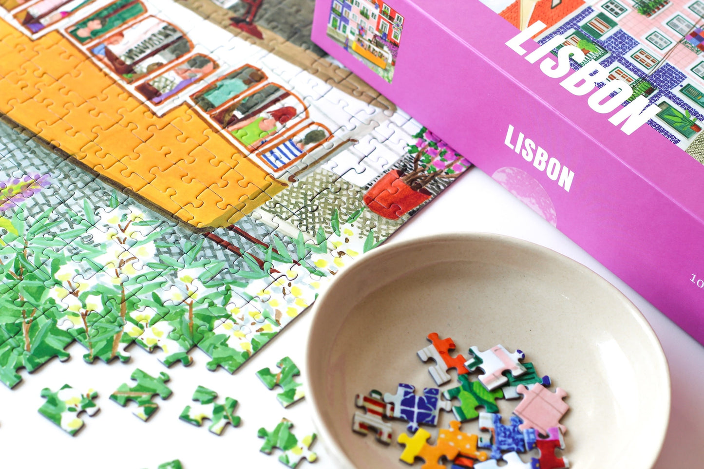 1000-Teile-Puzzle „Lisbon“ von Piecely – Illustration einer farbenfrohen Straßenszene in Lissabon mit gelber Straßenbahn und bunten Häusern, auf pinker Verpackung.