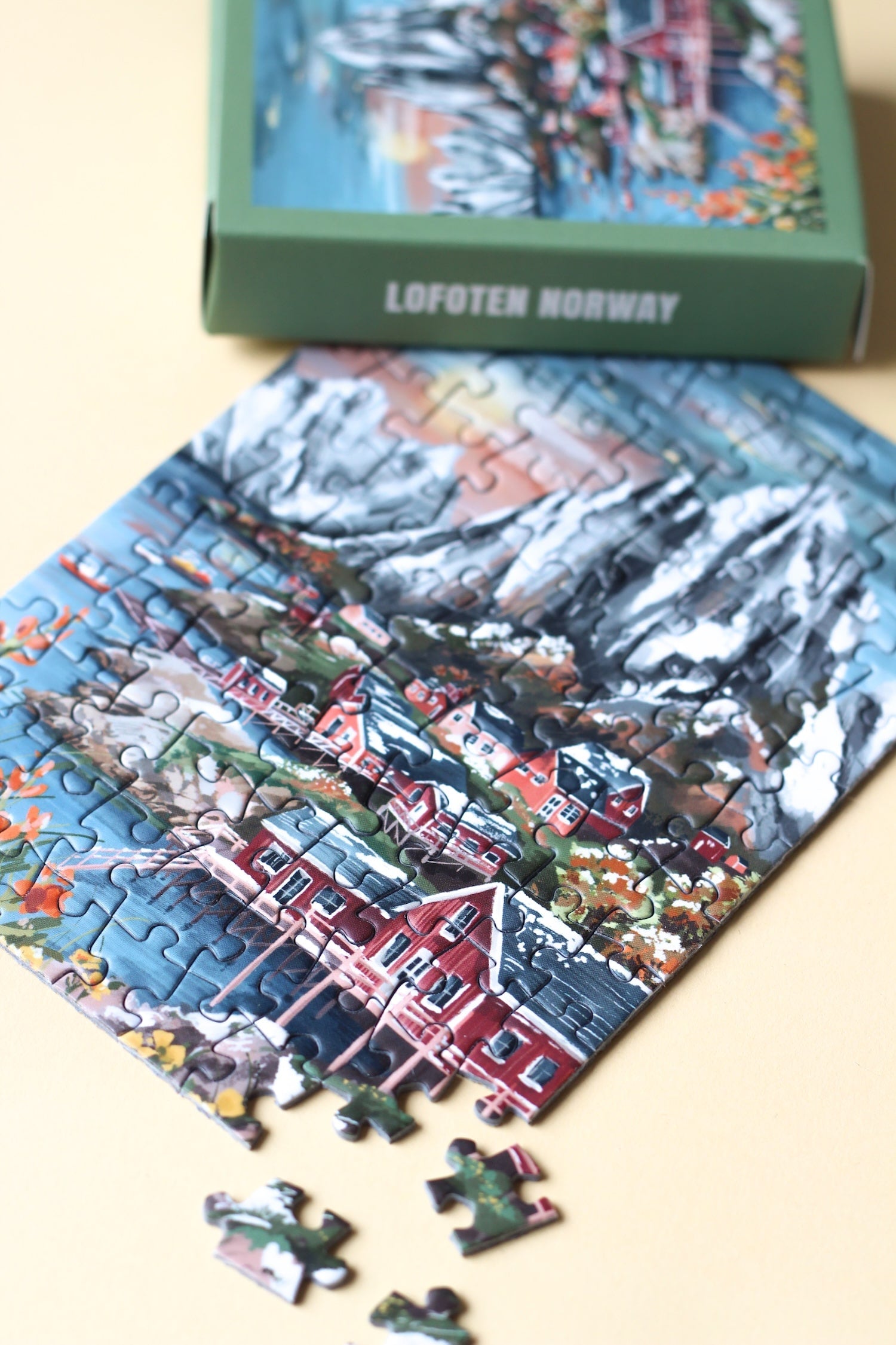 99-Teile-Mini-Puzzle „Lofoten Norway“ von Piecely – Illustration einer norwegischen Küstenlandschaft mit Bergen, roten Häusern und Meer, auf olivgrüner Verpackung.