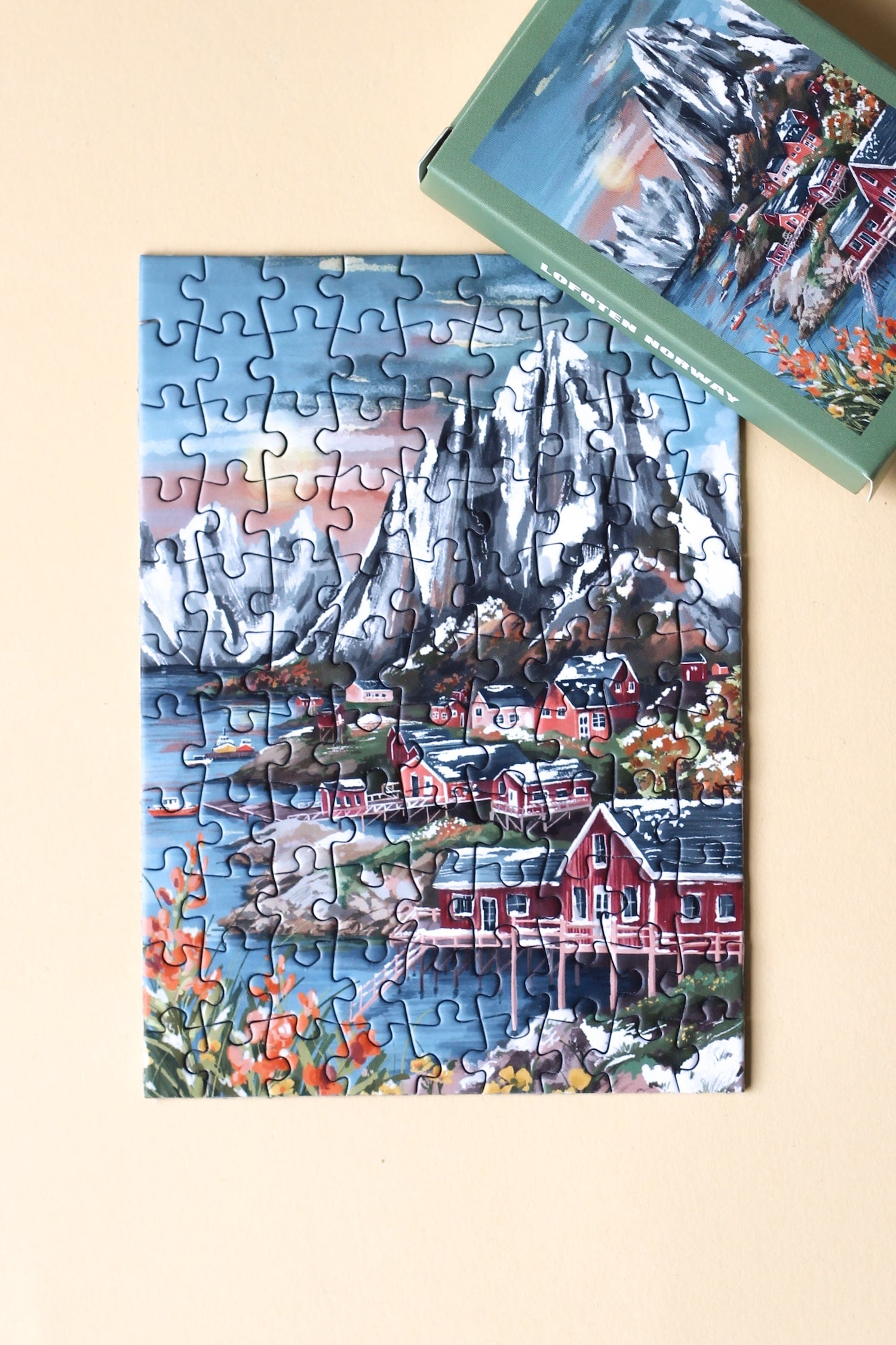 99-Teile-Mini-Puzzle „Lofoten Norway“ von Piecely – Illustration einer norwegischen Küstenlandschaft mit Bergen, roten Häusern und Meer, auf olivgrüner Verpackung.