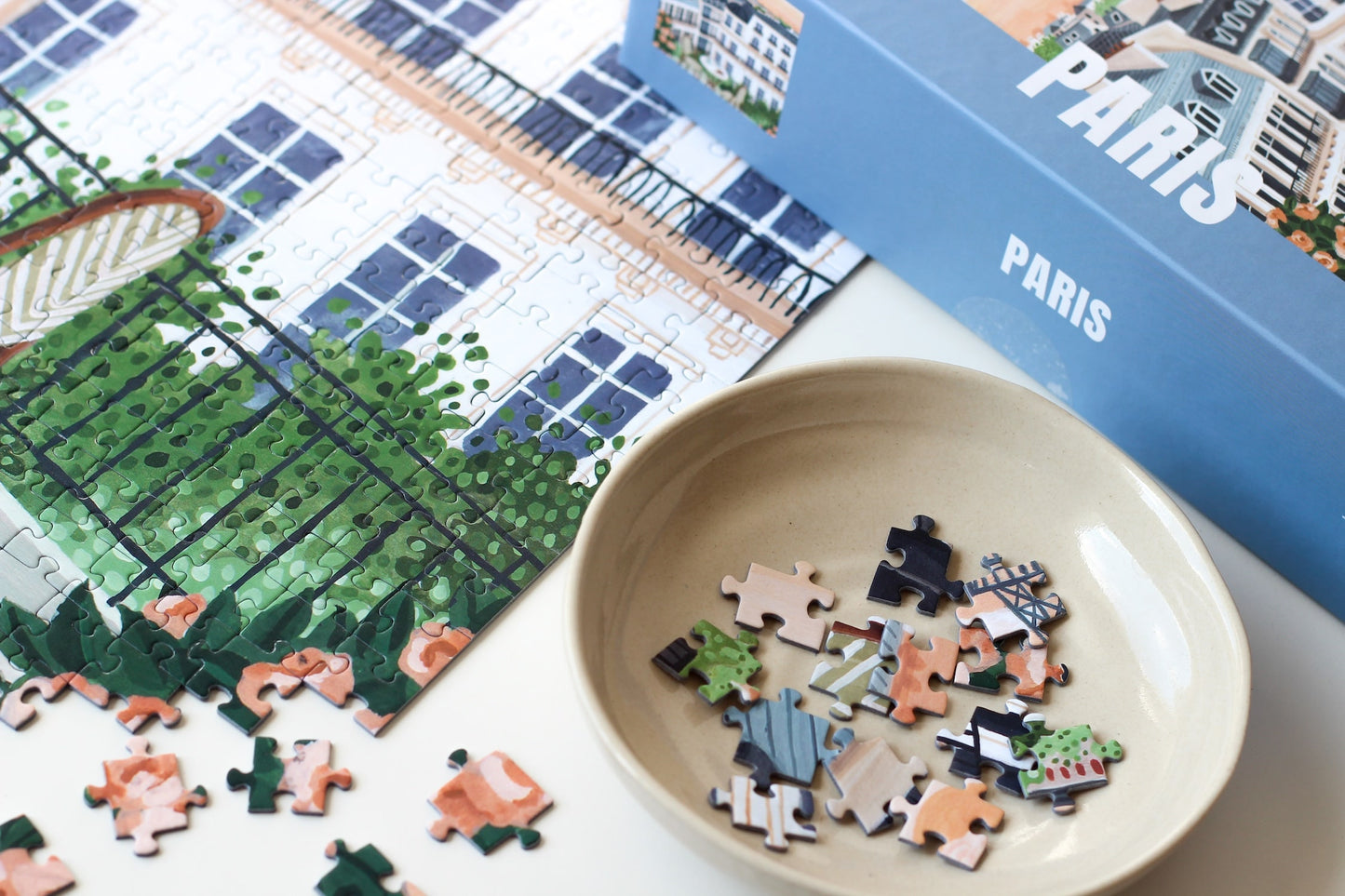 1000-Teile-Puzzle „Paris“ von Piecely – Illustration eines Pariser Balkons mit Frühstückstisch und Blick auf den Eiffelturm, auf hellblauer Verpackung.