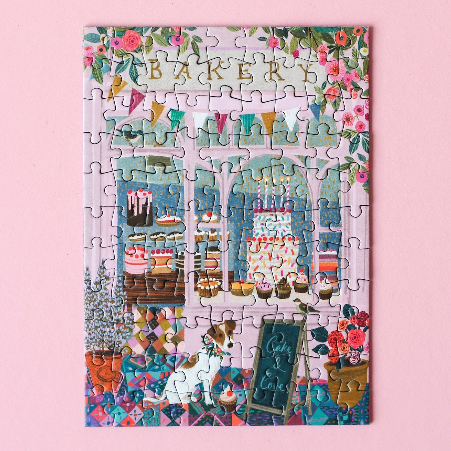 Plastikfreies Minipuzzle für Erwachsene, das eine rosa Bäckerei mit Torten und Cupcakes im Schaufenster und einen Hund vor der Tür zeigt.