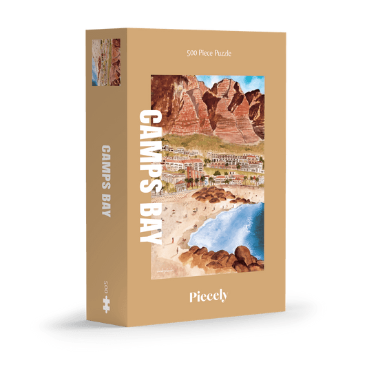 500-teiliges Puzzle für Erwachsene, das die Strandpromenade des Kapstädter Viertels Camps Bay zeigt. Im Vordergrund sind man Sandstrand und blaues Meer und im Hintergrund Häuserketten und die Berge.