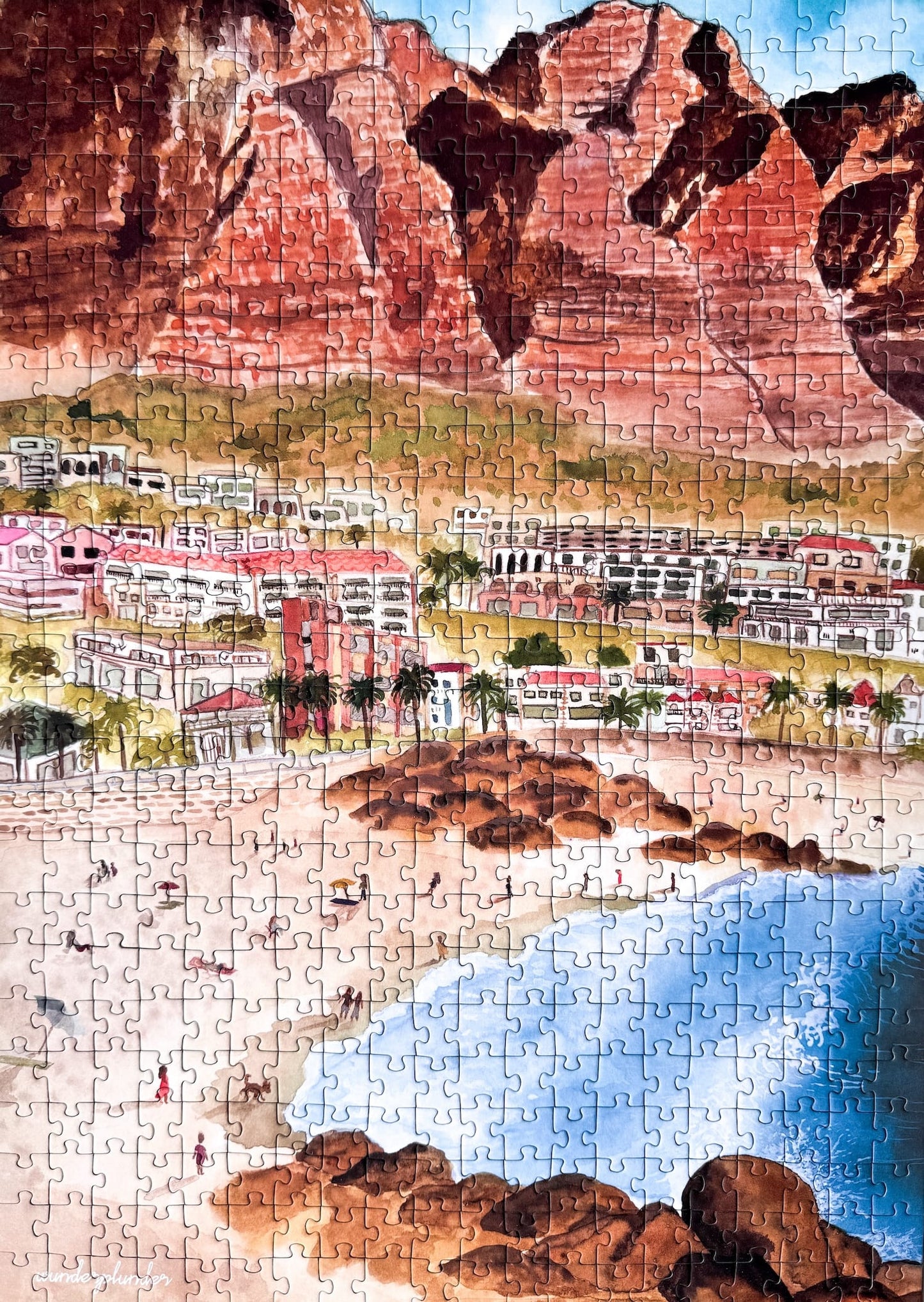 500-teiliges Puzzle für Erwachsene, das die Strandpromenade des Kapstädter Viertels Camps Bay zeigt. Im Vordergrund sind man Sandstrand und blaues Meer und im Hintergrund Häuserketten und die Berge.