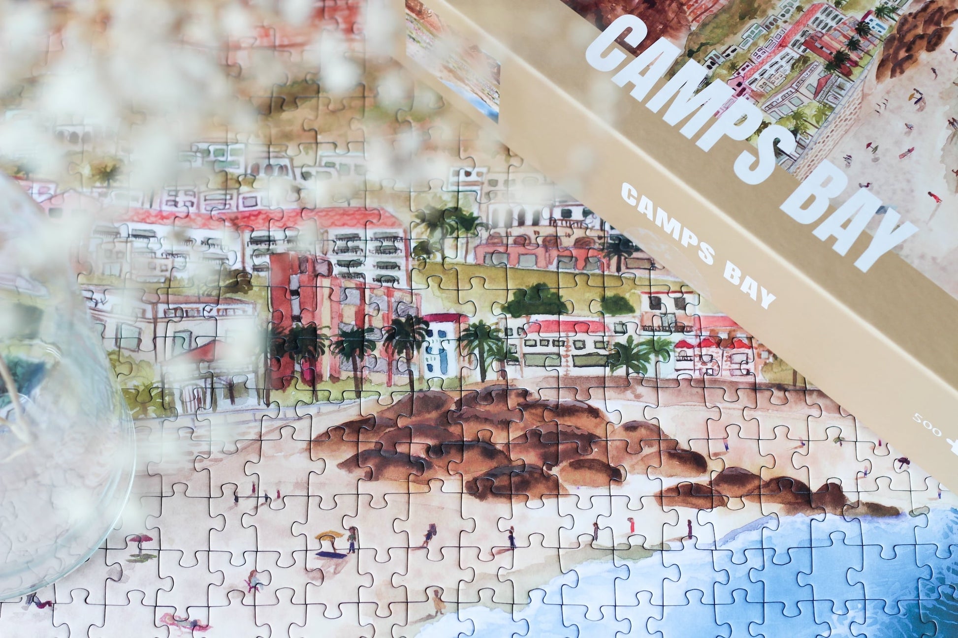 500-teiliges Puzzle für Erwachsene, das die Strandpromenade des Kapstädter Viertels Camps Bay zeigt. Im Vordergrund sind man Sandstrand und blaues Meer und im Hintergrund Häuserketten und die Berge.