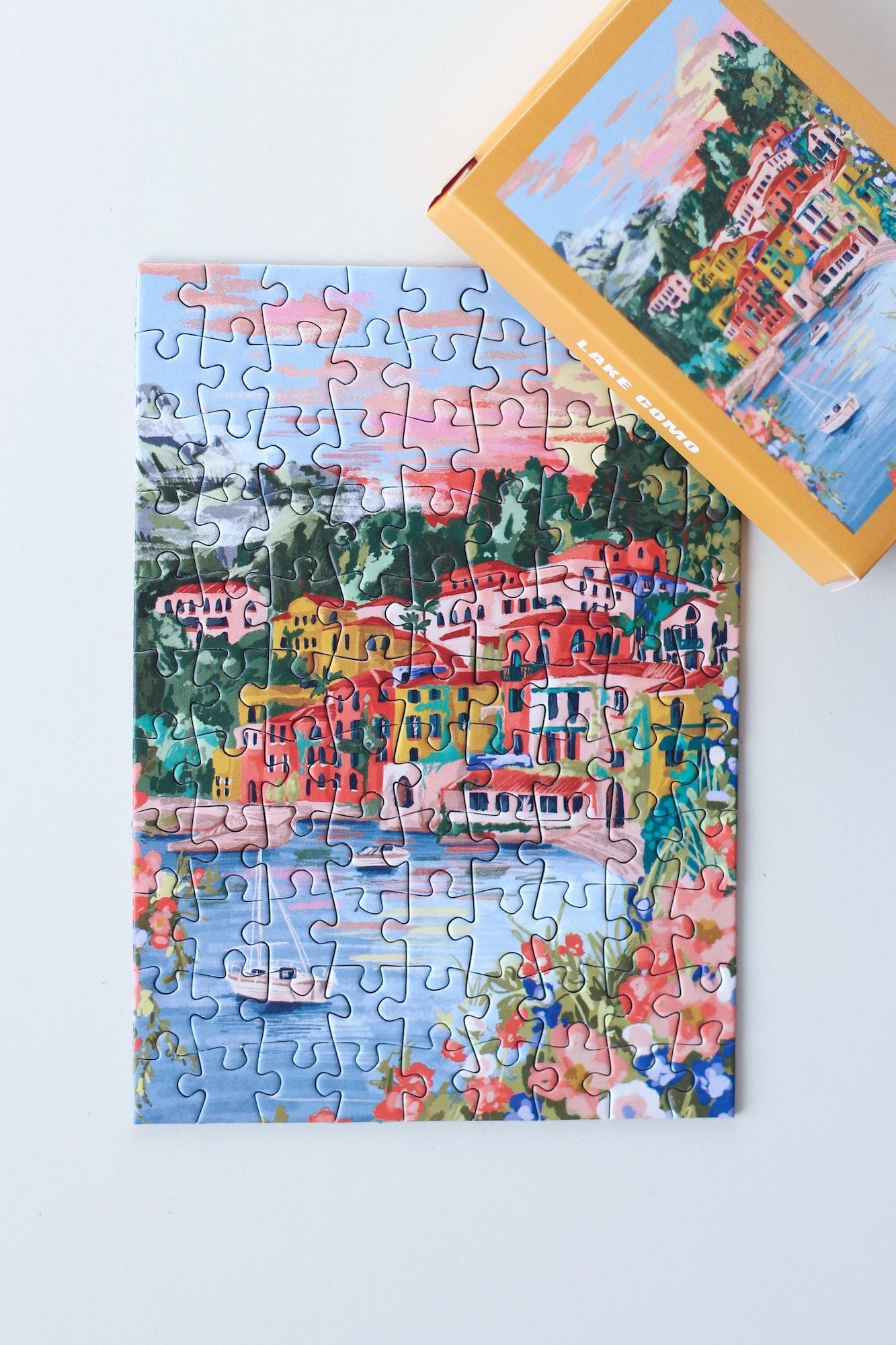 99-teiliges Minipuzzle für Erwachsene, das den Comer See in Varenna zeigt. In der Bucht befinden sich zwei Boote und die Sicht auf den See ist gesäumt von bunten Häusern.