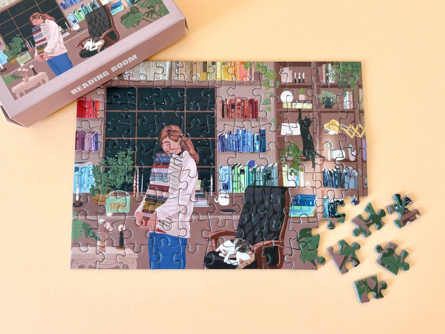 99-teiliges Minipuzzle für Erwachsene, das eine Frau mit einem Stapel Bücher in der Hand stehend vor ihrem gefüllten Bücherregal zeigt. Eine Katze schläft auf dem Stuhl neben ihr und eine andere klettert das Bücherregal hoch.
