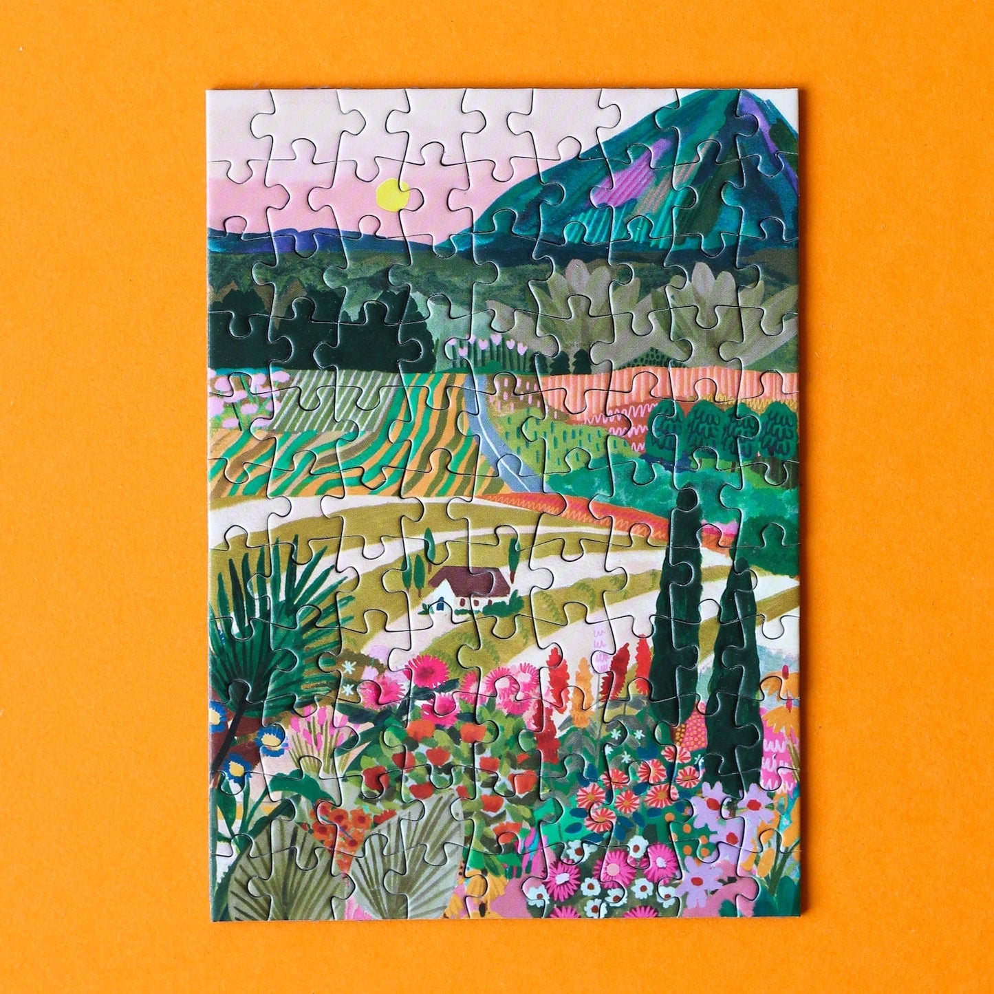 Plastikfreies Minipuzzle für Erwachsene, das die bunte Landschafts Apuliens in Italien zeigt. Zu sehen sind ein Haus, die Berge, bunte Blumen, Felder und die untergehende Sonne