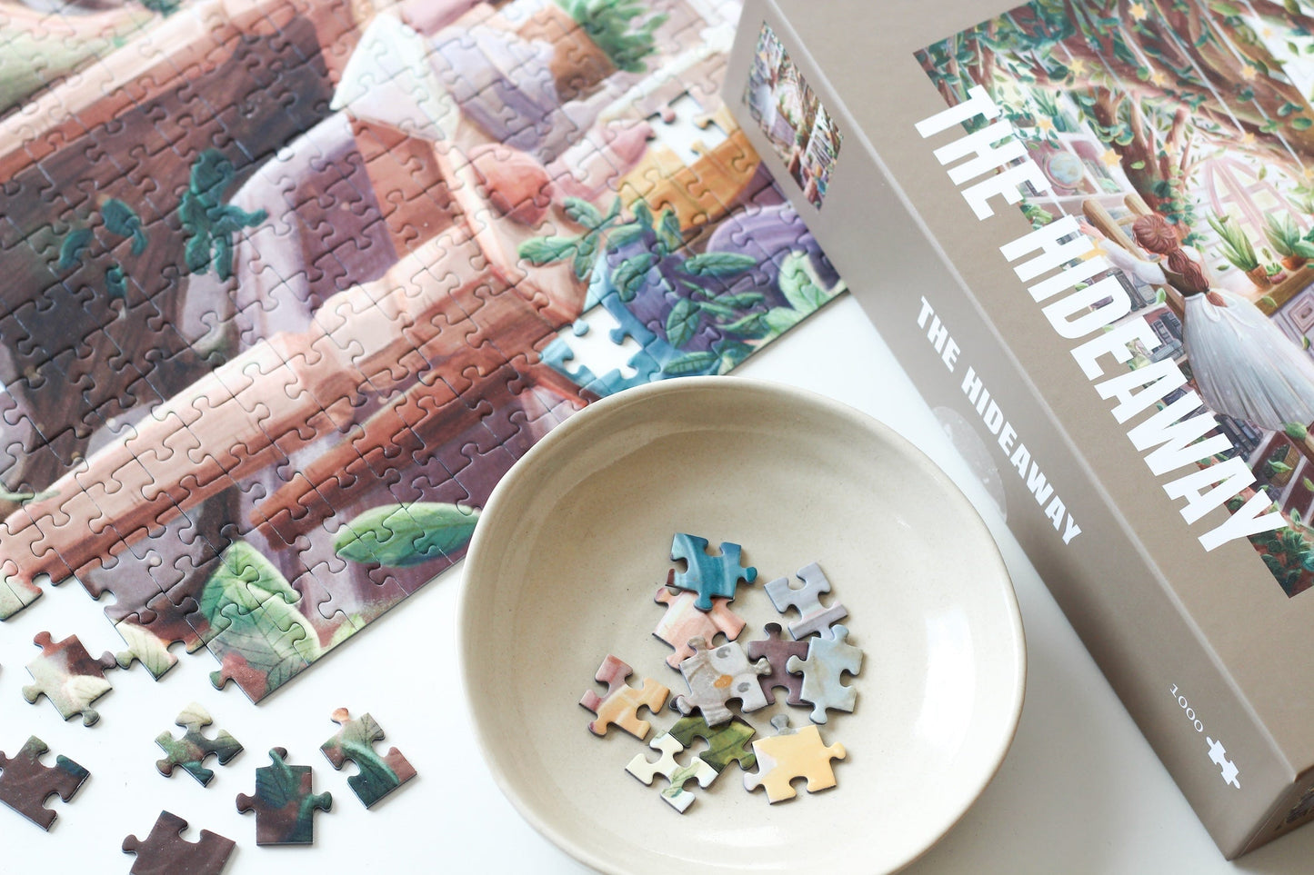 1000-teiliges Puzzle für Erwachsene, das eine Frau auf einer Leiter in einer verwunschenen Küche zeigt. Mitten in der Küche wächst ein Baum und die Regale sind gefüllt mit Büchern, Porzellan und Pflanzen.